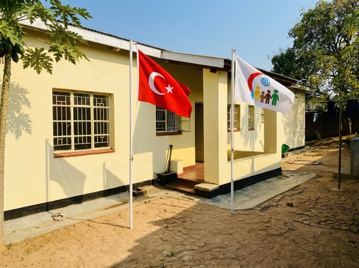 Temsili görsel: Mamaneso Yuvam'ın sarı badanalı evi; verandanın önünde Türk bayrağı ile Beş Yürek amblemli beyaz bayrak dalgalanıyor.