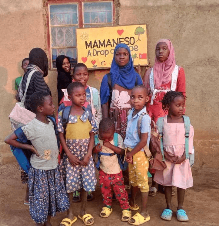 Yeni okul çantalarını takmış çocuklar, 'Mamaneso — A Drop of Hope' tabelasının önünde.